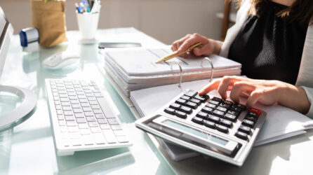 A woman sitting at a desk uses a calculator and references a binder on the desk