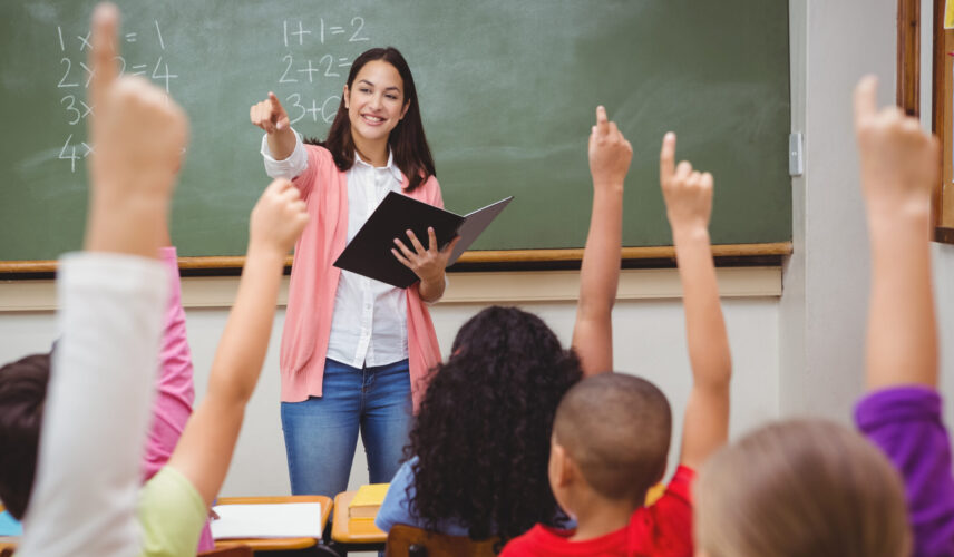 A teacher stands in front of a chalkboard and points to a room full of students with their hands up