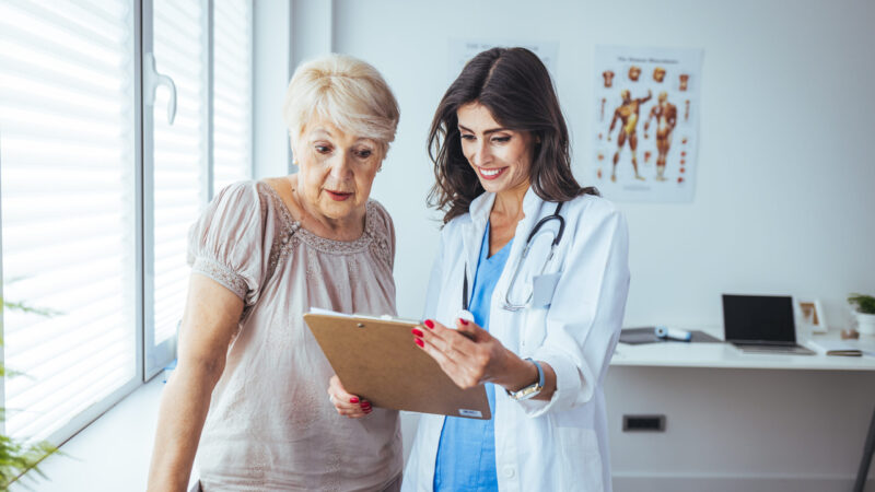A doctor holding a clipboard explains something to her patient, an older woman