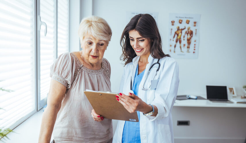A doctor holding a clipboard explains something to her patient, an older woman