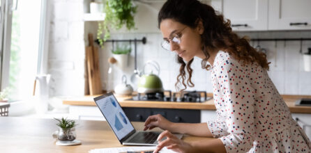 A woman standing at her kitchen counter using a laptop and calculator.