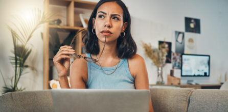 A young woman sits on a couch with her laptop computer in her lap, looking off into the distance and thinking.