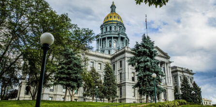 A view of the Colorado State Capitol Building framed by green trees on a mostly cloudy day.