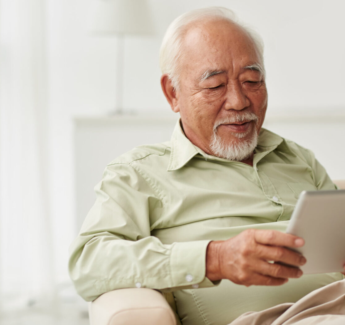 A white-haired Asian man sitting in a chair and reading on his tablet.