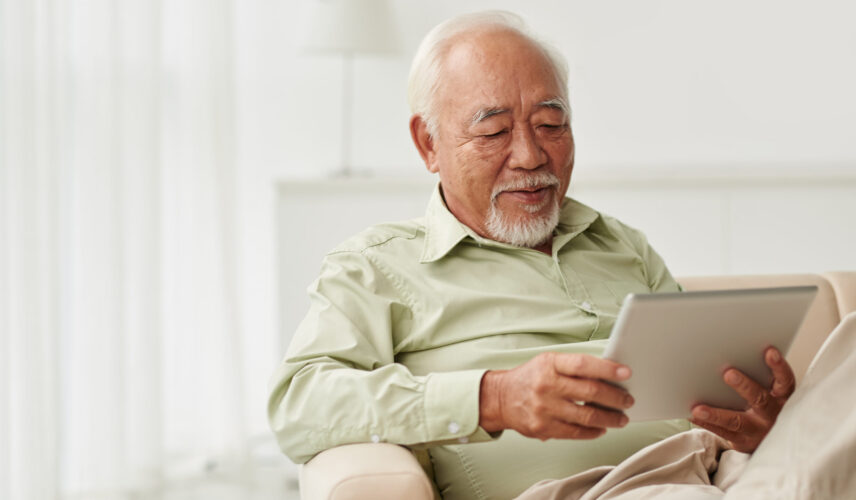 A white-haired Asian man sitting in a chair and reading on his tablet.